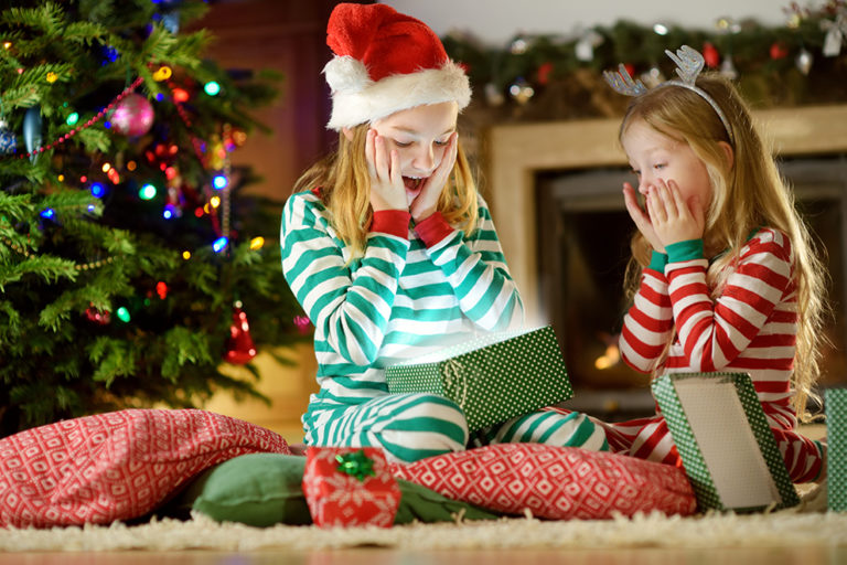 Happy little sisters wearing Christmas pajamas opening gift boxes by a fireplace in a cozy dark living room on Christmas eve. Celebrating Xmas at home. Happy little sisters wearing Christmas pajamas opening gift boxes by a fireplace in a cozy dark living room on Christmas eve. Celebrating Xmas at home.