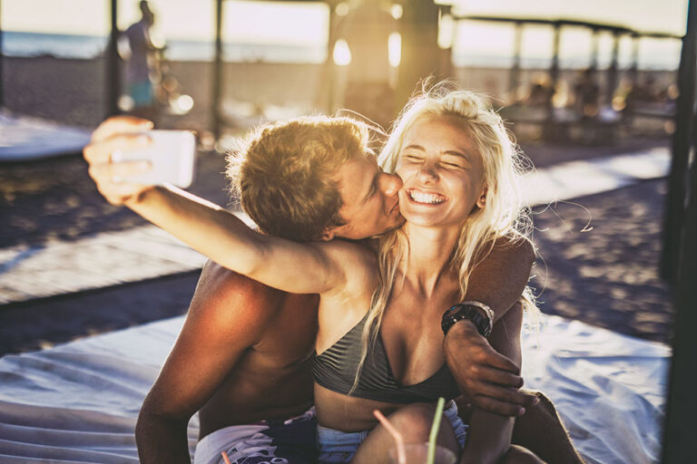 Young loving couple taking a selfie with mobile phone on the beach in summer day while man is kissing his girlfriend in the cheek.