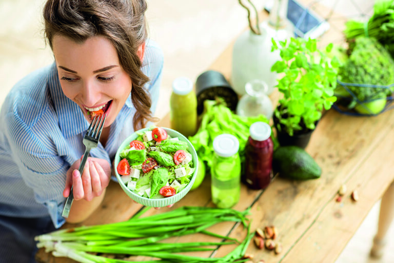 Young and happy woman eating healthy salad sitting on the table with green fresh ingredients indoors Young and happy woman eating healthy salad sitting on the table with green fresh ingredients indoors