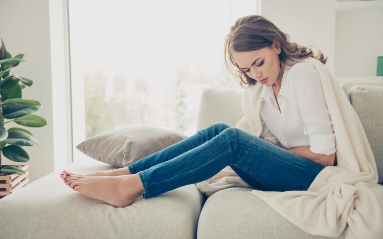 Sad, upset, unhappy woman holding hands on stomach suffering from abdominal pain with close eyes, having menstrual period, food poisoning, gastritis, diarrhea, feeling unwell sitting in livingroom Sad, upset, unhappy woman holding hands on stomach suffering from abdominal pain with close eyes, having menstrual period, food poisoning, gastritis, diarrhea, feeling unwell sitting in livingroom