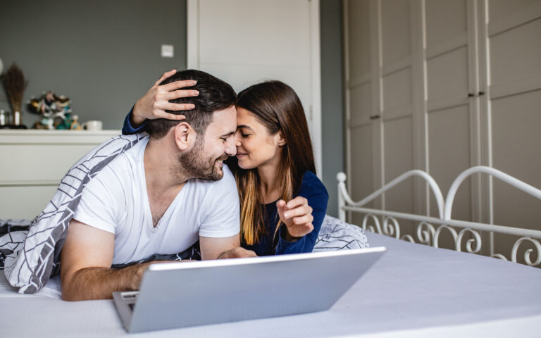 Young couple is laying in bed and they are using laptop and looking at each other Young couple is laying in bed and they are using laptop and looking at each other