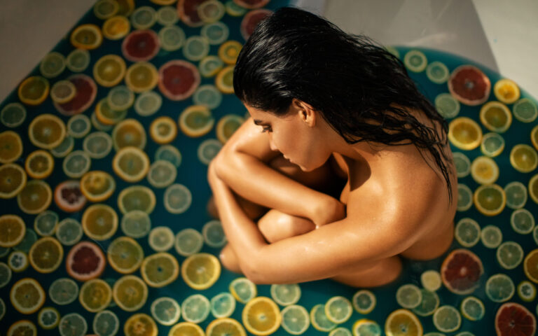 Portrait of beautiful fashion model posing in a bathtub filled with blue water and fresh fruit