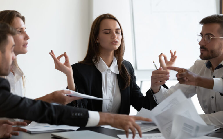 Mindful young business woman meditating, managing stress, ignoring annoyed arguing colleagues at workplace. Peaceful young female manager calming down during stressful working day at company office.