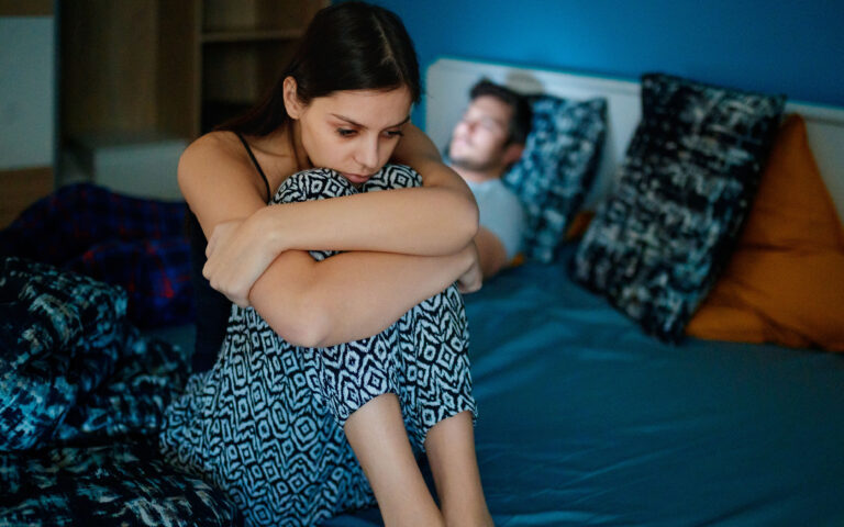 Sad girlfriend sitting on bed edge, holding her legs after fight, her partner is lying in bed in background, his face is lit by smart phone Sad girlfriend sitting on bed edge, holding her legs after fight, her partner is lying in bed in background, his face is lit by smart phone