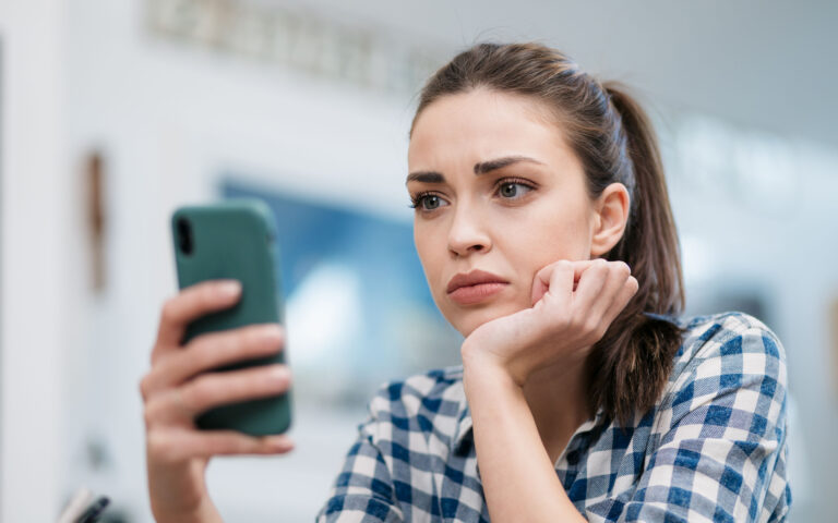 Close up of a depressed Caucasian young woman sitting at home, reading some bad news on her smart phone, feeling sad and worried