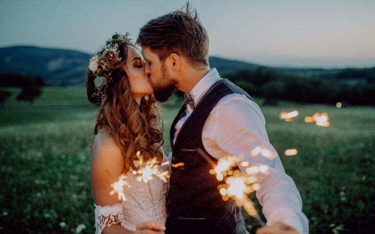 Beautiful young bride and groom on a meadow in the evening, holding sparklers.
