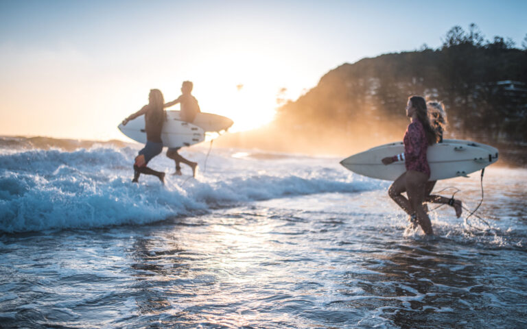 Four friends, surfers, running into the water early in the morning with surfboards in their hands. Sun is rising in back.