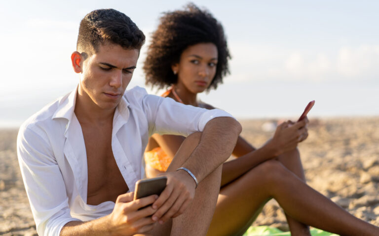 A girl of African descent is jealous and watches her boyfriend hide his cell phone messages, while theyâre on vacation, sitting on the beach. A girl of African descent is jealous and watches her boyfriend hide his cell phone messages, while theyâre on vacation, sitting on the beach.