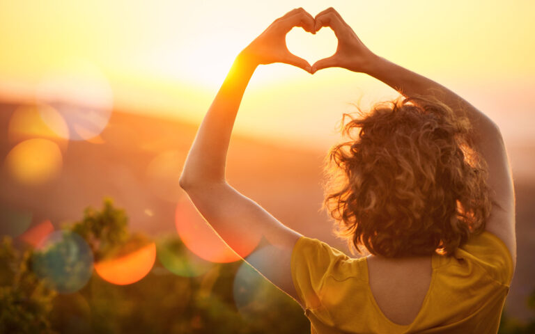 Rearview shot of an unidentifiable woman making a heart shape with her hands over a sunset landscape Rearview shot of an unidentifiable woman making a heart shape with her hands over a sunset landscape