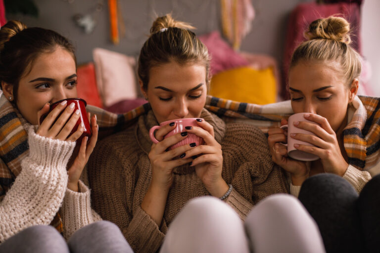 Front view of three pretty female friends in winter clothes drinking hot beverages during cold day inside a room. Front view of three pretty female friends in winter clothes drinking hot beverages during cold day inside a room.