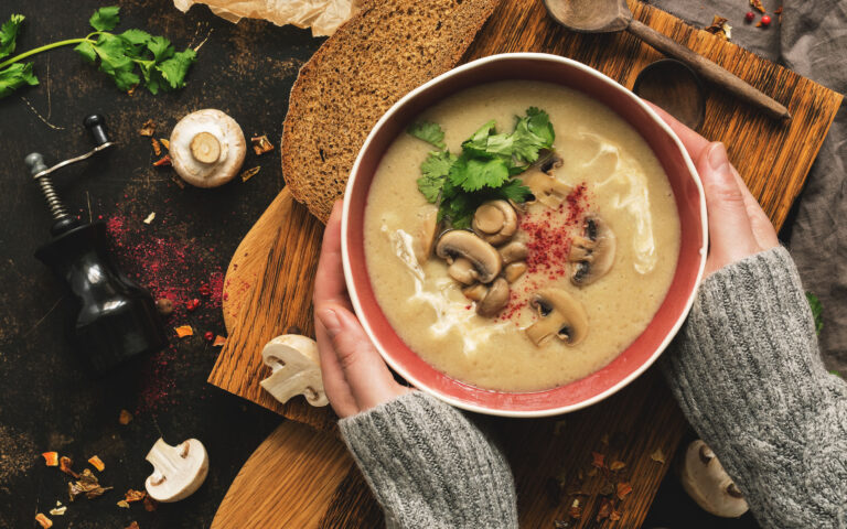 Bowl of mushroom cream soup in woman hands in a woolen sweater. Rustic dark background, top view, flat lay. Winter warming soup on vintage cutting boards Bowl of mushroom cream soup in woman hands in a woolen sweater. Rustic dark background, top view, flat lay. Winter warming soup on vintage cutting boards