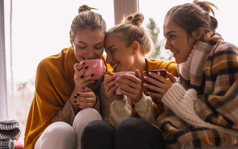 Close-up of three happy close female friends in woolen clothes sitting by the window while sharing hot beverage time. Close-up of three happy close female friends in woolen clothes sitting by the window while sharing hot beverage time.