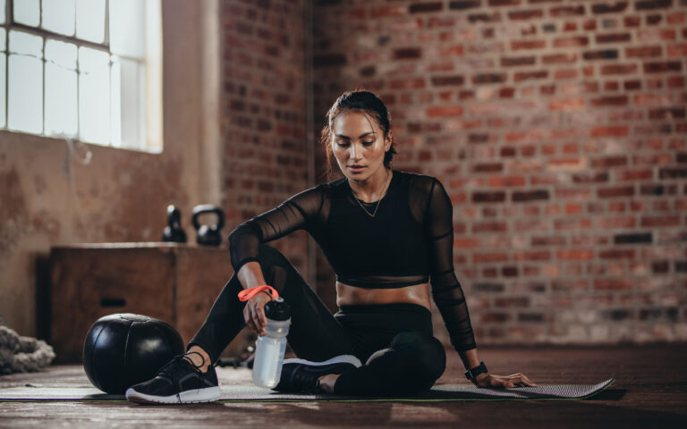Portrait of a fit young woman taking a break in the gym. Female sitting on fitness mat with a water bottle looking down. Portrait of a fit young woman taking a break in the gym. Female sitting on fitness mat with a water bottle looking down.