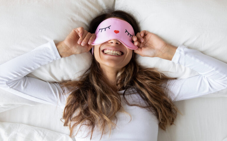 Top view close up happy woman wearing funny sleeping mask enjoying morning in bedroom, lying in comfortable bed on soft pillow, beautiful girl with healthy toothy smile stretching in the morning