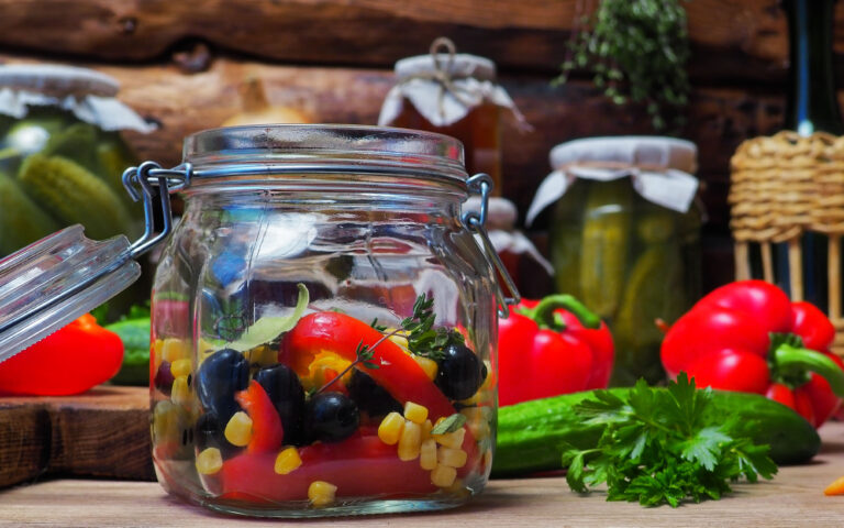 Cooking of homemade preserves with fresh vegetable: cucumbers and bell peppers, carrot and sweetcorn, leek and olives. Fresh and dry herbs and seasonings on kitchen table amid ready veggie preserves