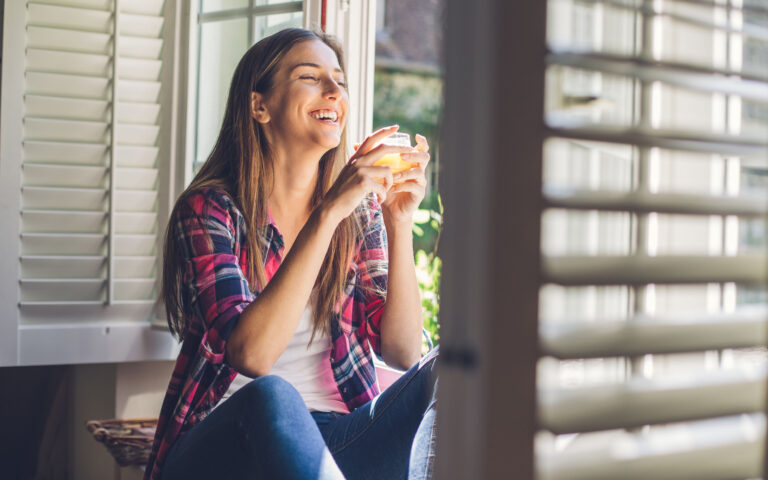 Smiling young woman holding a glass with orange juice, sitting on the windowsill in a country villa. Smiling young woman holding a glass with orange juice, sitting on the windowsill in a country villa.