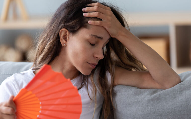 Close up view millennial woman sitting on couch exhausted by hot weather closed eyes feels unwell dying of heat holding in hand orange fan using it for reducing too hot temperature indoors concept