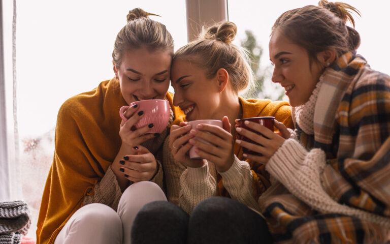 Close-up of three happy close female friends in woolen clothes sitting by the window while sharing hot beverage time.