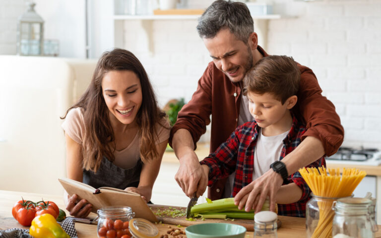 Mother, father, son chopping vegetables on kitchen. Salad preparation for dinner or lunch. Father helping boy child chopping celery. Toothy smiling mother telling recipe from cookbook. Happy family