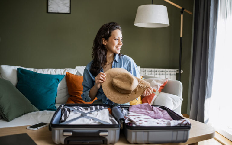 Young woman sitting on the couch and packing suitcase. She is holding a straw hat in her hands Young woman sitting on the couch and packing suitcase. She is holding a straw hat in her hands