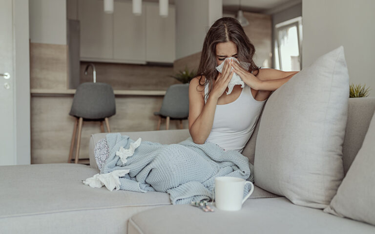 Sick Woman Covered With a Blanket Lying in Bed With High Fever and a Flu, Resting at Living Room. She Is Exhausted and Suffering From Flu. Sick Woman With Runny Nose Lying in Bed. Girl Suffering From Cold Lying in Bed With Tissue Blowing Her Nose While Sitting on the Sofa