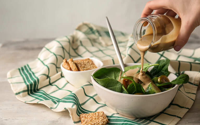 Pouring of tasty tahini from jar onto fresh vegetables in bowl