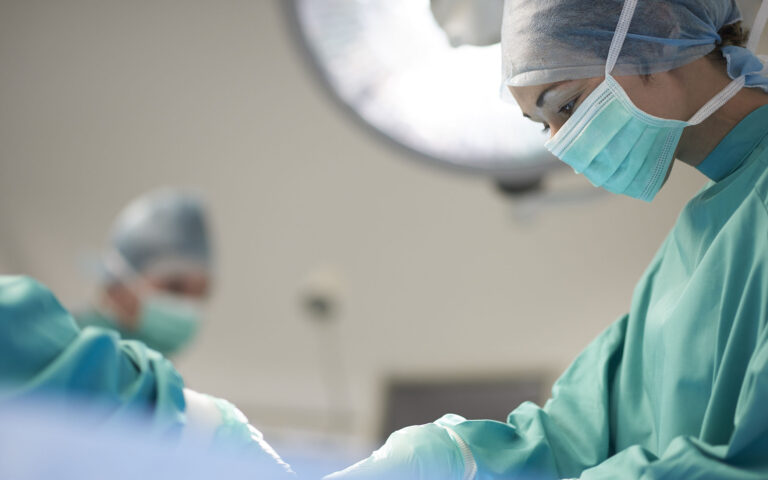 a female operating nurse stands over a patient at the operating table and looks down to what she is doing in the operation. In the background the anaesthetist is looking over from his monitors . both surgeons or nurses are female in the foreground .