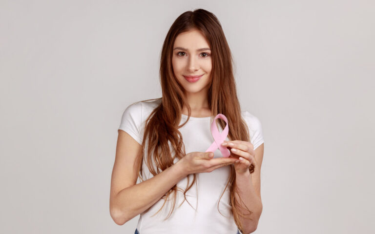 Women's health care, timely diagnosis. Happy smiling optimistic woman holding pink ribbon, symbol of breast cancer awareness, wearing white T-shirt. Indoor studio shot isolated on gray background.