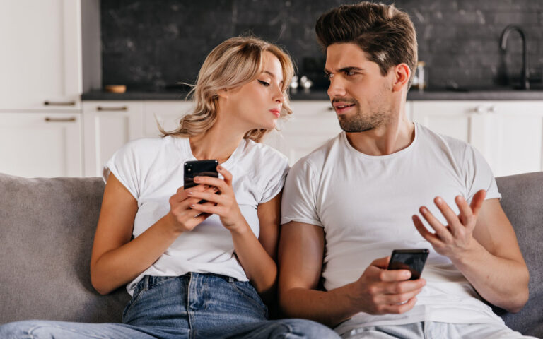Curly woman looking at husband's phone. Appealing caucasian girl sitting on sofa with smartphone.